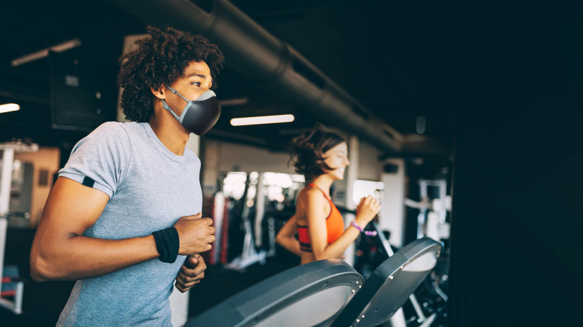 一名戴著黑色 LG PuriCare 穿戴式空氣清新機的非裔美國男子正在健身室的跑步機上跑步。他看起來呼吸順暢，神態自若。