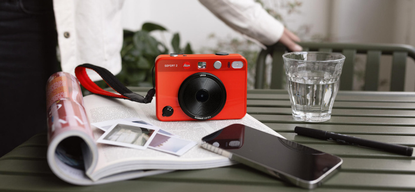 The Leica Sofort 2 red with prints and a mobile phone on a table.